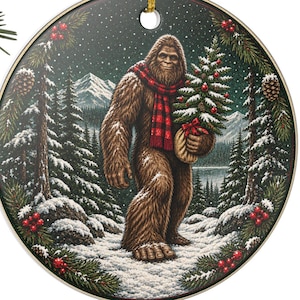 May include: A round ceramic ornament featuring a brown Bigfoot creature wearing a red and black buffalo plaid scarf, carrying a small decorated Christmas tree in a snowy forest landscape with mountains, pine branches, and red berries.