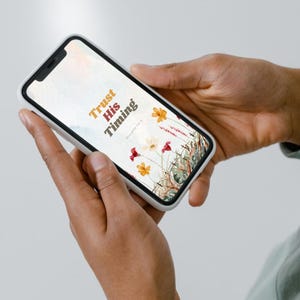 Può includere: Uno smartphone che mostra una grafica digitale con il testo Trust His Timing e Proverbs 3:5, caratterizzata da un'illustrazione ad acquerello di fiori di campo colorati nelle tonalità del rosso, arancione e giallo su sfondo chiaro.