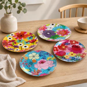 May include: Four round ceramic plates featuring vibrant, multi-colored floral patterns. One plate has a yellow background, one is blue, one is pink, and one is white. Each plate displays a unique arrangement of stylized flowers in shades of red, orange, purple, and green.