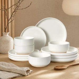 May include: A dinnerware set consisting of white ceramic plates and bowls with a smooth finish and exposed light brown clay bases. The collection includes stacked round dinner plates, salad plates, and deep bowls arranged on a wooden surface.