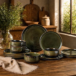 May include: A set of dark green ceramic dinnerware featuring a reactive glaze finish with subtle brown edges. The collection includes large dinner plates, smaller salad plates, and deep bowls arranged on a wooden surface with a beige linen napkin.