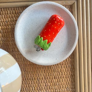 Pode incluir: Um isqueiro cilíndrico com uma capa vermelha em forma de morango, com pequenos pontos amarelos e uma parte superior de folhas verdes. O isqueiro está posicionado dentro de um prato de cerâmica redondo, salpicado e de cor esbranquiçada.