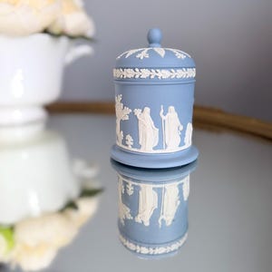 Puede incluir: Un tarro cilíndrico de color azul claro con tapa a juego, que presenta adornos en relieve blanco de figuras clásicas y follaje. El recipiente de cerámica con acabado mate muestra intrincados detalles blancos sobre el fondo azul.