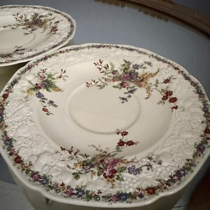 Può includere: Due piattini in ceramica color bianco sporco con un bordo in rilievo raffigurante frutta e viti. Il centro di ogni piattino è decorato con un delicato motivo floreale multicolore nei toni del rosso, viola, blu e giallo, con bordi smerlati.