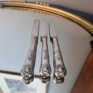 May include: Three silver-toned metal knives with ornate, embossed shell-like patterns on the handles. Each knife features a smooth, straight blade and a decorative, flared end with detailed scrollwork and a prominent fan-shaped motif.