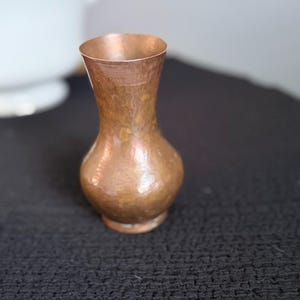 Könnte beinhalten: Eine kleine Vase aus gehämmertem Kupfer mit bauchigem Körper und ausgestelltem Rand. Die Metalloberfläche weist eine ausgeprägte, texturierte Struktur mit Farbvariationen auf, die von hellen Kupfertönen bis zu dunkleren, oxidierten Brauntönen reichen.