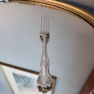 May include: A silver-toned metal fork featuring a four-tined head and a decorative handle with an embossed shell-like motif at the base. The handle displays intricate scrollwork and a central floral-inspired design, reflecting a classic metallic finish.