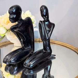 May include: Two glossy black sculptures of kneeling figures with smooth, elongated forms. One figure is positioned slightly forward with hands near the chest, while the other is positioned behind, both featuring minimalist, featureless facial structures.