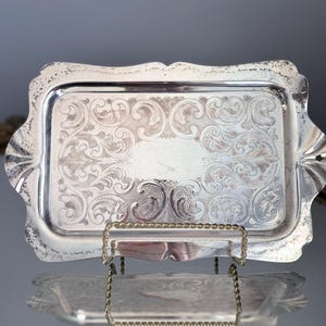 Peut inclure: Un plateau rectangulaire en métal argenté orné d'un motif gravé de volutes sur toute sa surface. Le plateau présente un bord festonné avec des détails décoratifs et une finition polie, présenté verticalement sur un support en fil doré.