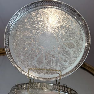 Peut inclure: Un plateau de service rond en métal argenté présentant un motif floral et des volutes en relief sur toute sa surface. Le plateau possède un bord ajouré en forme de treillis et une zone centrale lisse et polie pour la présentation.