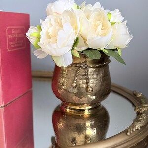 Puede incluir: Un macetero de latón martillado con un detalle decorativo de borde estilo cuerda, lleno de un ramo de flores de peonía artificiales de color blanco y crema. La superficie metálica del macetero refleja la luz, mostrando un acabado texturizado.