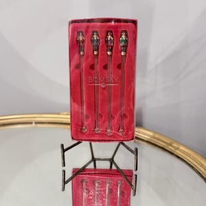 Peut inclure: Un ensemble de quatre agitateurs à cocktail en verre transparent avec des embouts décoratifs en forme de casse-noisette rouges, bleus, violets et verts. Ils sont présentés dans une boîte rectangulaire rouge marquée BOMBAY en blanc.