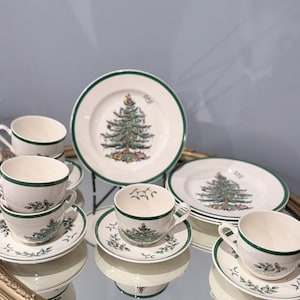 Puede incluir: Un juego de tazas de té y platillos de cerámica color crema con un borde verde y una ilustración de árbol de Navidad. Los platillos están decorados con motivos de acebo y bayas, mientras que las tazas muestran un pequeño diseño de árbol.
