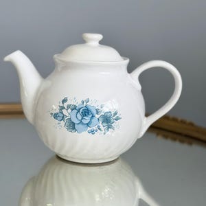 May include: A white ceramic teapot featuring a delicate blue floral pattern of roses and leaves on the front. The teapot has a rounded body with subtle vertical ribbing, a matching lid with a small knob, a curved spout, and a handle.