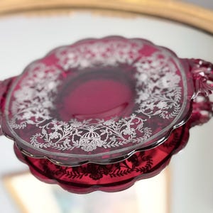 Puede incluir: Un plato de servicio redondo de cristal rojo intenso con bordes festoneados y dos asas ornamentadas. La superficie presenta un detallado patrón floral plateado grabado que rodea el centro, creando un borde decorativo sobre el cristal rubí.