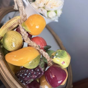 Puede incluir: Un racimo de frutas decorativo para colgar que presenta réplicas de cerámica de un plátano, naranja, pera, manzana y uvas. Las frutas están atadas con una cuerda de yute gruesa de color natural, con acabados brillantes en tonos amarillos, rojos y morados.