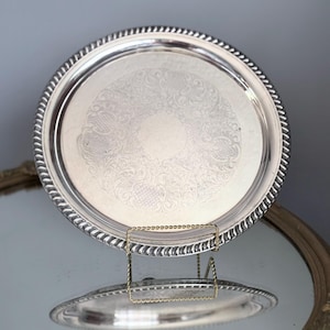 May include: A round silver-toned serving tray featuring a polished finish and a decorative rope-style border. The centre of the tray displays an intricate, etched floral pattern surrounding a smooth, circular blank space in the middle.
