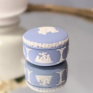 Puede incluir: Un joyero redondo de cerámica azul claro con tapa extraíble. El exterior presenta adornos en relieve de color blanco que representan figuras clásicas y guirnaldas florales. La superficie es mate y el diseño es uniforme en todo el contorno.