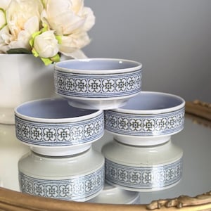 Puede incluir: Tres cuencos de cerámica blanca con un patrón floral geométrico repetido en azul y gris en el exterior. Los cuencos tienen un interior liso de color azul claro y una base corta elevada, presentados en una disposición apilada sobre una superficie.