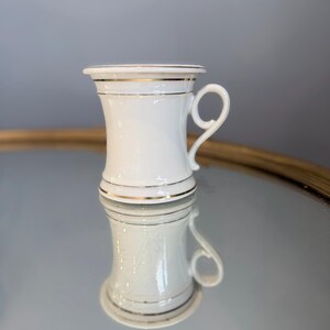 May include: A white porcelain mug featuring a flared rim and a curved handle. The mug is decorated with thin, horizontal gold bands around the top, middle, and base, providing a simple and clean aesthetic for this classic drinking vessel.