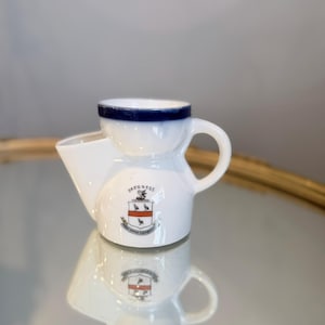 May include: A white ceramic shaving mug with a dark blue rim and a side handle. The front features a printed crest with the text SKEGNESS above it and a Latin motto below. The mug has a wide, angled spout on the left side for pouring water.