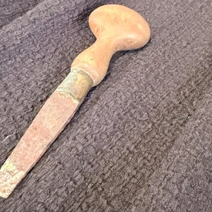 Op de afbeelding: Een handgereedschap met een gladde, afgeronde houten handgreep bevestigd aan een plat, taps toelopend metalen blad. Het metalen gedeelte vertoont tekenen van oxidatie en slijtage met een doffe afwerking, terwijl de houten handgreep lichtbruin is.