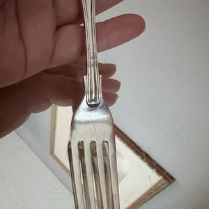 May include: A silver-toned metal fork featuring an ornate, embossed floral pattern on the handle. The utensil has four long, slender tines and a polished finish, with a decorative design element at the base of the neck where it meets the handle.