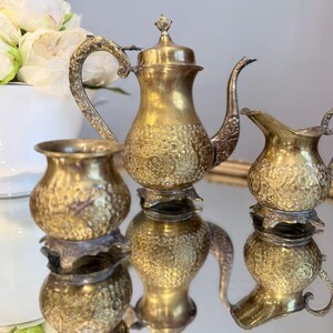 May include: A three-piece tea set featuring a teapot, a creamer, and a sugar bowl. Each piece is crafted from brass with an intricate embossed floral pattern, a curved handle, and a pedestal base. The teapot includes a lid with a small finial.