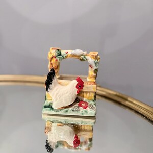 May include: A small ceramic figurine featuring a white chicken with a black tail and red comb standing in front of a yellow and brown fence structure. The fence is decorated with green foliage and small red flowers, with a white bird perched on top.