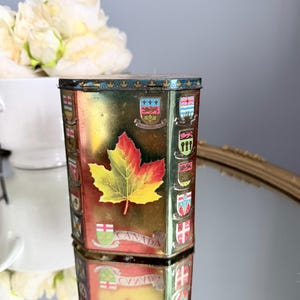 Peut inclure: Une boîte en métal hexagonale ornée d'un grand motif de feuille d'érable aux tons vifs jaune et rouge au centre. La boîte est décorée de divers blasons provinciaux et territoriaux colorés et du mot CANADA imprimé en lettres capitales.