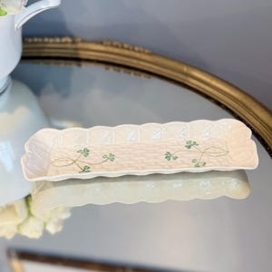 May include: A rectangular off-white ceramic tray with a scalloped edge and a textured basket-weave pattern on the base. The interior surface features two delicate green shamrock motifs painted on the center of the tray.
