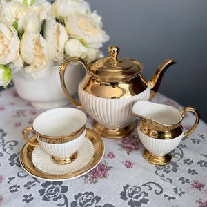Puede incluir: Un juego de té que incluye una tetera, una jarra de leche y una taza con plato. Cada pieza está hecha de cerámica blanca con detalles acanalados verticales y acentos dorados pulidos en las tapas, bordes, asas y bases de los recipientes.