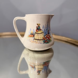 May include: A cream-coloured ceramic pitcher featuring a colourful illustration of a person in a yellow dress standing in a garden with blue flowers and orange blossoms. The pitcher has a curved handle and a wide spout, with a smooth glazed finish.