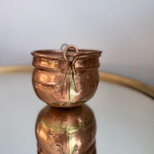 May include: A small, round copper pot with a metallic finish and a visible seam running down the side. The pot features a curved handle attachment on the rim, showing signs of oxidation and wear, with a warm, reflective reddish-brown surface.