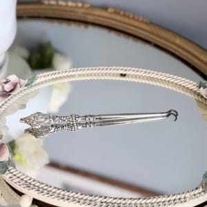 Puede incluir: Un gancho para botones de plata con un mango ornamentado y en relieve, con intrincados motivos de volutas y detalles florales. El eje largo y delgado termina en un gancho de metal curvado para abotonar, con un acabado metálico pulido.