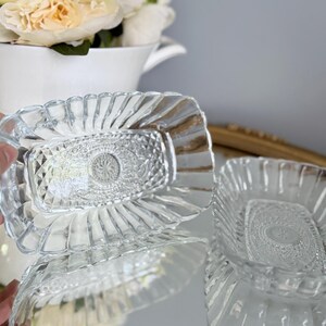 May include: Two rectangular clear glass dishes featuring a pressed pattern with a central circular medallion, diamond-shaped facets, and a scalloped, fluted rim. The glass is transparent and reflective, showcasing intricate geometric detailing.