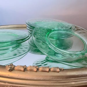 Puede incluir: Una pila de platos de vidrio verde claro translúcido con un patrón texturizado de hendiduras circulares a lo largo del borde. Los platos están dispuestos sobre una superficie reflectante, creando un efecto visual en capas con el vidrio verde.