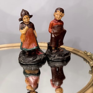May include: Two painted figurines of children standing on dark, textured bases. The left figure wears a dark brown hat, a dark top, and a reddish-brown apron. The right figure wears a dark cap, a reddish-orange long-sleeved top, and dark brown trousers.
