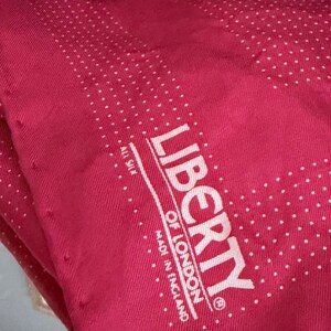 May include: A deep pink silk scarf featuring a white polka dot pattern. The fabric is printed with the white text LIBERTY OF LONDON, ALL SILK, and MADE IN ENGLAND, framed by a series of small white dots arranged in a rectangular border design.