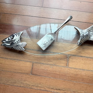 May include: A clear oval glass serving platter featuring a metallic silver fish head at one end and a matching metallic silver fish tail at the other. A matching metallic silver serving spatula with a textured handle rests on the glass surface.
