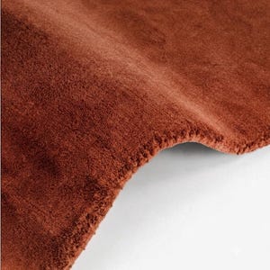 Puede incluir: Una alfombra de felpa de color sólido en un tono naranja terracota profundo y cálido. El textil presenta una superficie densa de pelo corto con una textura suave y aterciopelada. La alfombra se muestra doblada, revelando el material grueso.