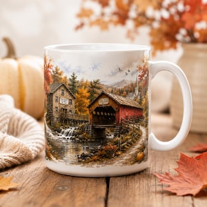 May include: A white ceramic mug featuring a detailed illustration of a rustic stone mill and a red covered bridge over a flowing stream. The scene includes autumn foliage, a small waterfall, and a church steeple in the distance. The text on the mill reads Pine Creek Old Mill Est 1885.