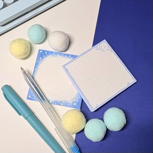 May include: Two square sticky note pads with celestial designs, one featuring a grid pattern with a moon and stars, the other with a dotted center and star border. Included are a blue pen, a clear gel pen, and six soft, fuzzy pom-pom balls in pastel colors.