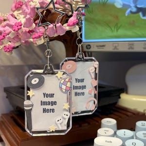 Può includere: Due portachiavi con cornice portafoto rettangolare in acrilico con chiusure a forma di stella. Ogni cornice presenta un inserto bianco con il testo 'Your Image Here' circondato da piccoli adesivi decorativi tra cui un microfono, un disco in vinile, una palla da discoteca e cuffie.