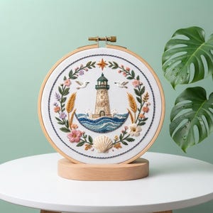 Op de afbeelding: Een ronde borduurring met een wit stoffen paneel met een handgeborduurde vuurtorenscène. Het ontwerp toont een centrale vuurtoren omringd door blauwe oceaangolven, twee witte vogels, een zeeschelp en een kleurrijke bloemenkrans.
