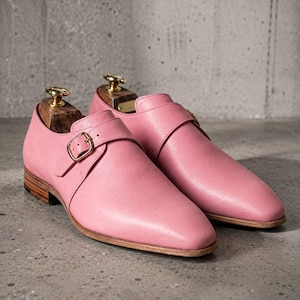 May include: A pair of pink leather monk strap shoes featuring a single gold-toned metal buckle closure, a smooth rounded toe, and a stacked wooden heel. Each shoe contains a wooden shoe tree with a gold-toned metal knob handle inserted inside.