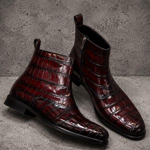 Könnte beinhalten: Ein Paar dunkelburgunderfarbene Stiefeletten mit glänzender Krokodilprägung. Die Stiefel haben eine spitz zulaufende Form, einen seitlichen Reißverschluss, eine Anziehschlaufe an der Ferse sowie einen schwarzen Absatz und eine Sohle.