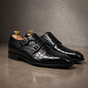 May include: A pair of black double monk strap shoes featuring a textured crocodile-embossed leather finish. Each shoe is equipped with two polished gold-tone metal buckles and a light-coloured wooden shoe tree inserted into the heel area.