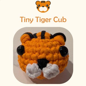 May include: A crocheted tiger cub head featuring bright orange yarn, black yarn stripes, two large black plastic eyes, and a white yarn muzzle with a black nose. The text Tiny Tiger Cub is displayed above the object on a white background.