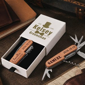 May include: Two multi-tool pocket knives with wooden handles, each engraved with a name and the date 2026.10.10. One knife is inside a white sliding gift box printed with a top hat, bow tie, and the text Kelsey Groomsman Est 1989.